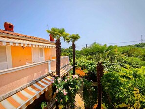 A building with a tiled roof, a balcony with a striped awning, and a garden with various plants and flowers.