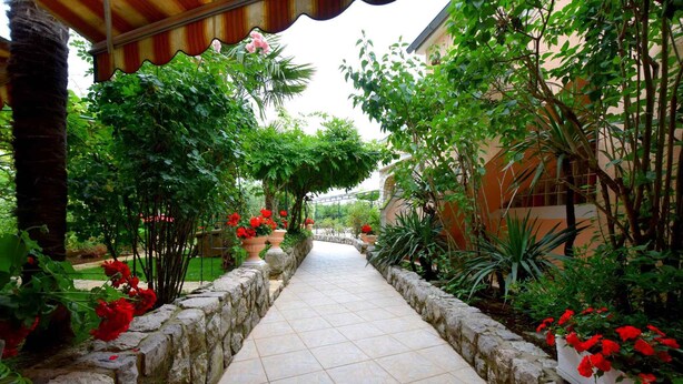 Un vialetto in pietra costeggiato da vegetazione lussureggiante e fiori rossi, che conduce a un edificio con tetto in tegole.