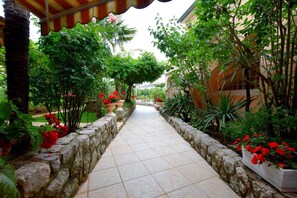 A stone pathway flanked by lush greenery and red flowers, leading to a building with a tiled roof. - Holiday home with terrace (Malinska)