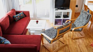 Una sala de estar con un sofá rojo, un sillón estampado, una mesita blanca con una planta y un televisor sobre un mueble blanco.
