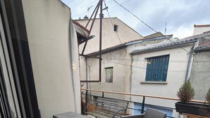 Un balcón con mobiliario de mimbre, barandilla metálica y vistas a edificios cercanos.