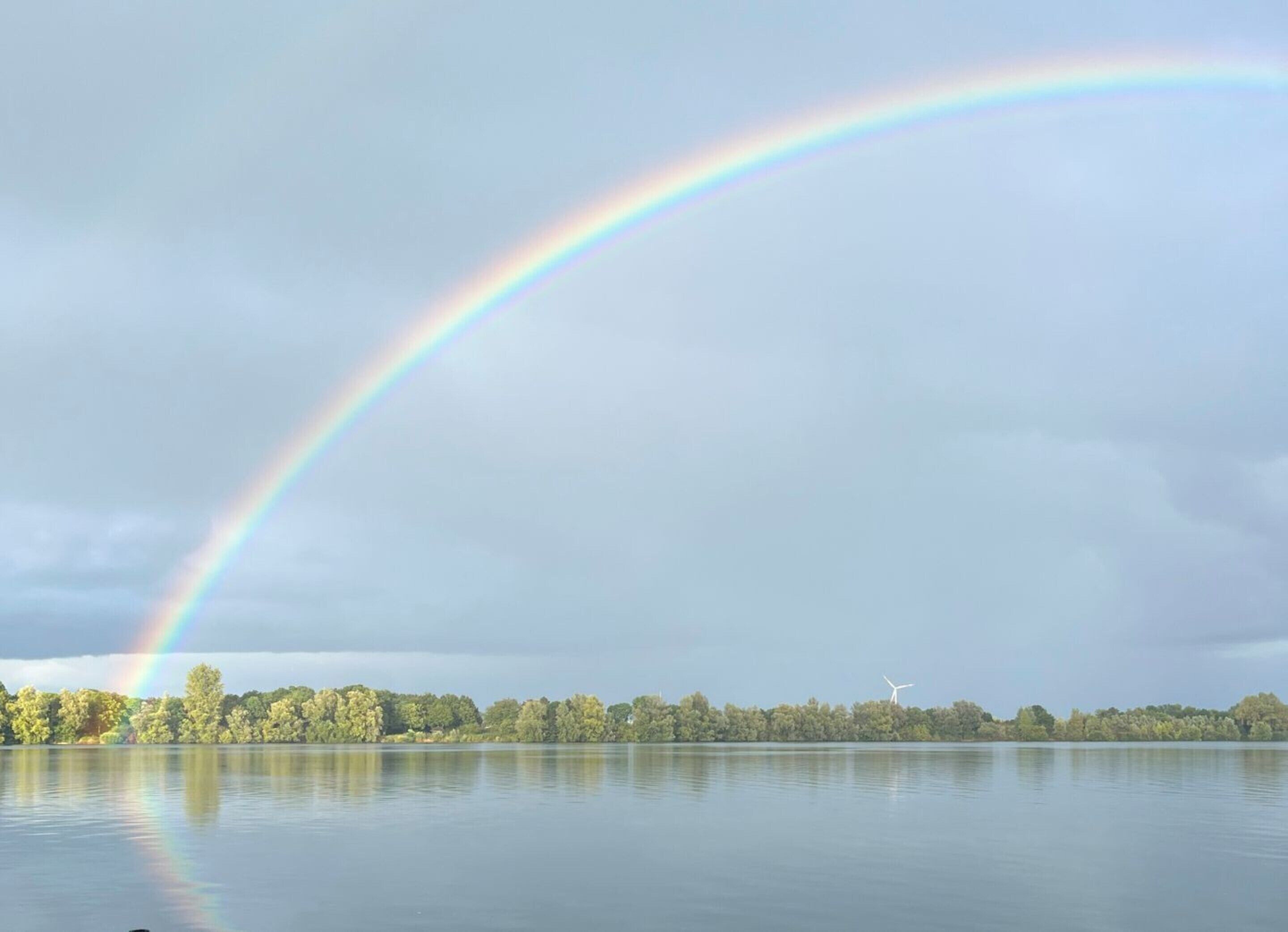 Un lac paisible, surmonté d’un arc-en-ciel, entouré d’arbres et avec une éolienne visible au loin.