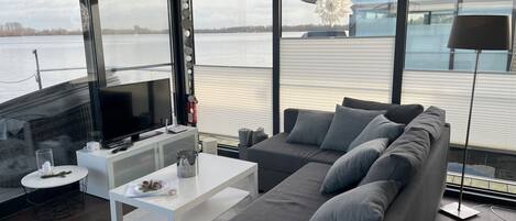 A modern living room with a grey sofa, white coffee table, and a flat-screen TV. A large window offers a view of a body of water and a building.