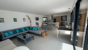 A modern living room with a grey sectional sofa, a coffee table, and a dining area with chairs. There is a kitchen area with a bar and a refrigerator. - VILLA Sea View with beautiful swimming pool and enclosed garden, seaside SOUTH CORSICA   (Pianottoli-Caldarello)