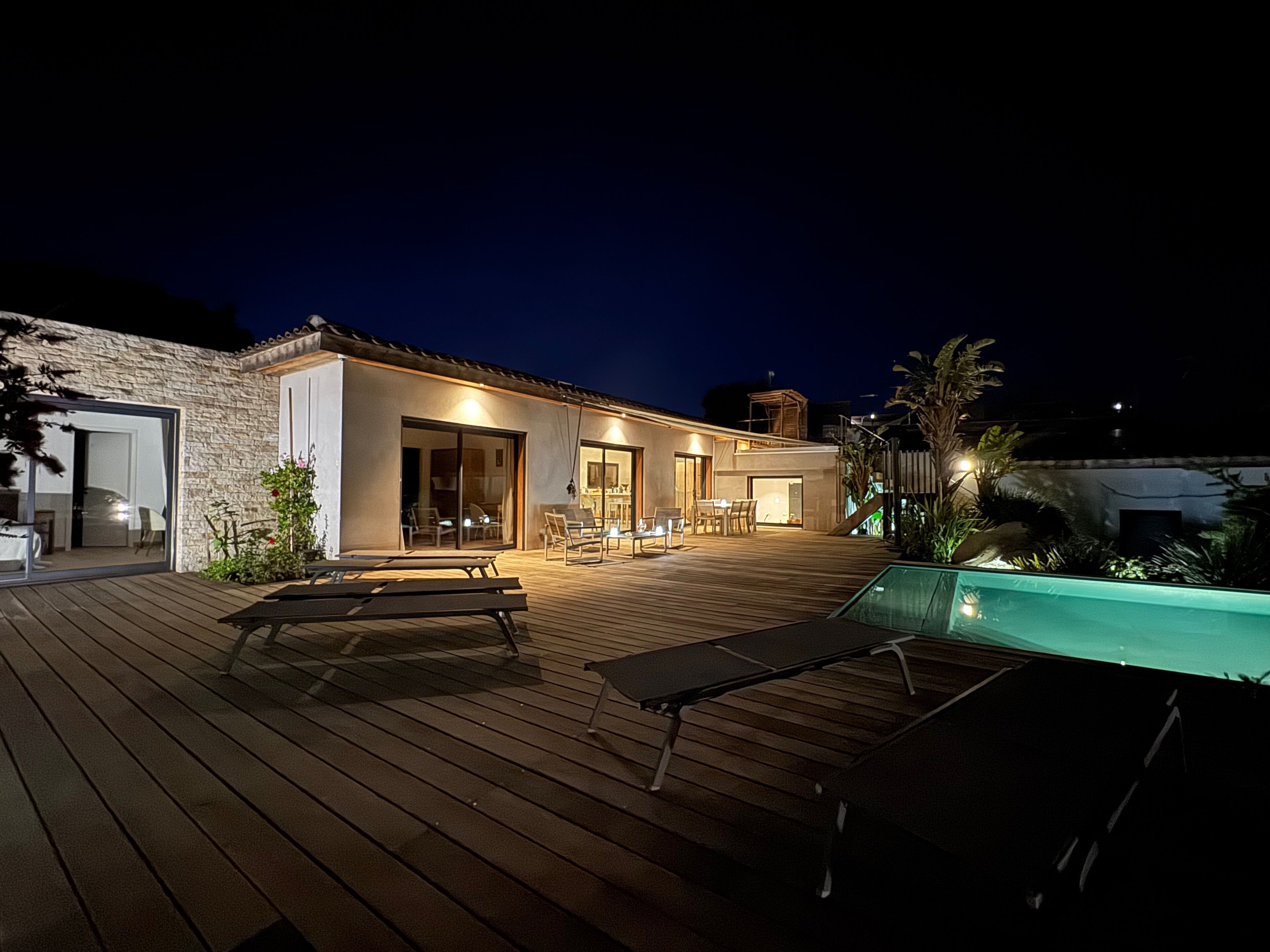 Ein modernes Haus mit einer Holzterrasse, Sitzgelegenheiten im Freien und einem Swimmingpool.