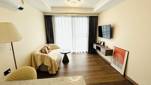 Ein Hotelzimmer mit Sofa, einem kleinen runden Tisch, einem an der Wand befestigten Fernseher und einem Fenster mit Vorhängen.