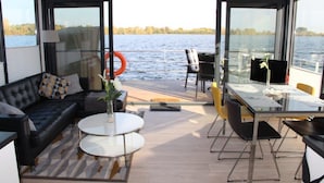 Un espace de vie moderne avec un canapé en cuir noir, deux tables rondes et une table à manger avec des chaises. Une grande fenêtre offre une vue sur un plan d’eau et des arbres.