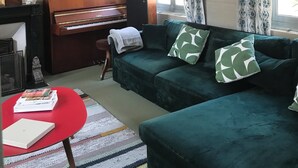 Un salon avec un canapé vert, une table rouge, un piano et une grande fenêtre ornée de rideaux à motifs.