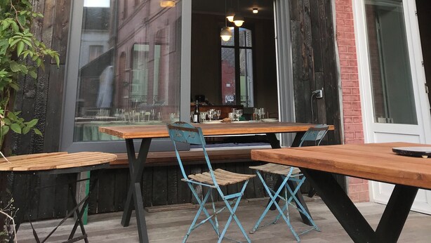 Un espace extérieur aménagé avec des tables en bois et des chaises pliantes bleues, une pergola en bois et, au fond, un bâtiment en briques.