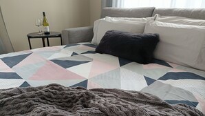 Un letto rifatto con cura, con una coperta a motivi geometrici e una coperta lavorata a maglia scura, più un tavolino con una bottiglia e dei bicchieri.
