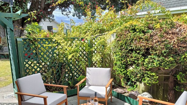 Un patio con mobili in legno e un tavolo rotondo, immerso nel verde e circondato da una recinzione in legno.