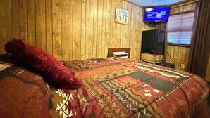 A room with wooden panel walls, a bed with a patterned bedspread, a television on the wall, and a window with blinds. - Short Branch Motel (Pahrump)