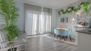 Una sala de estar moderna con un sofá, una mesa de comedor con sillas, una zona de cocina con microondas y un ventanal con cortinas.
