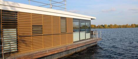 A modern houseboat with a wooden exterior, large windows, and a platform extending over the water.
