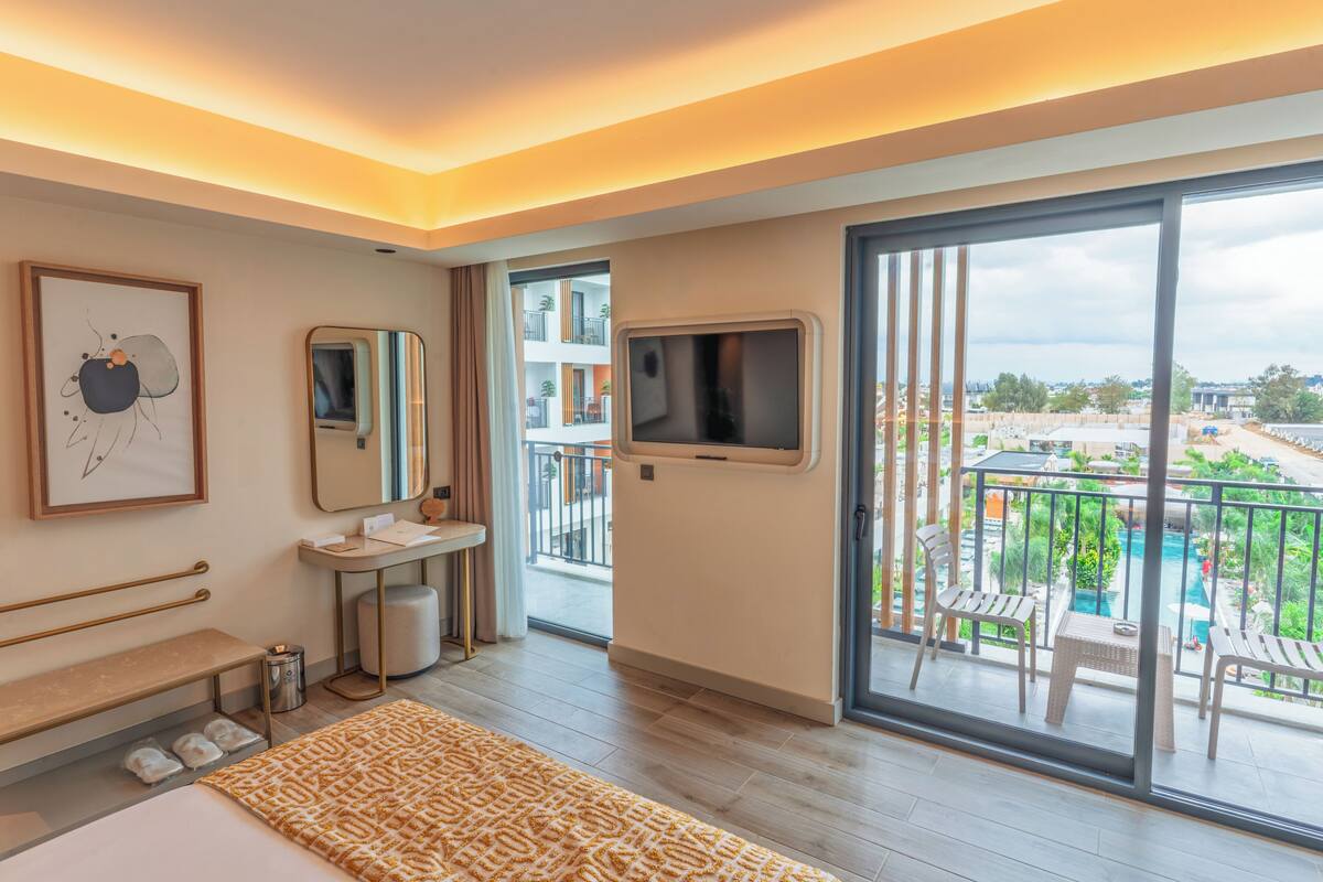 A modern hotel room with a large bed, a bathroom with a vanity, a TV, and a balcony with a pool view.