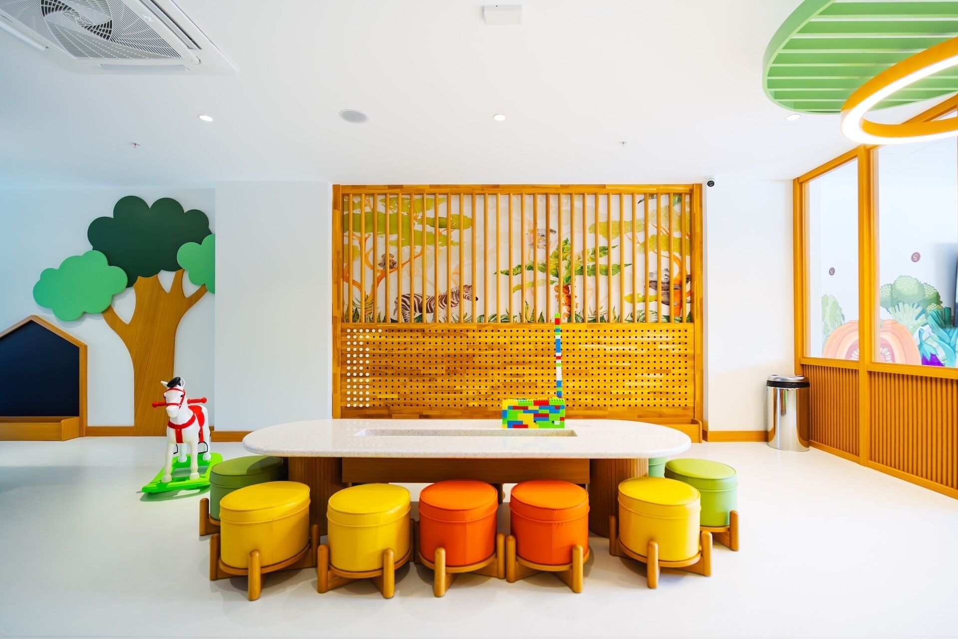 Un área de juegos para niños con una mesa grande, taburetes coloridos y una valla decorativa de madera con ilustraciones de animales.