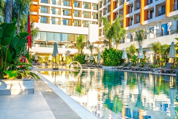 Una piscina con sillas de descanso y sombrillas, rodeada por un edificio de hotel con múltiples balcones y zonas de estar al aire libre.