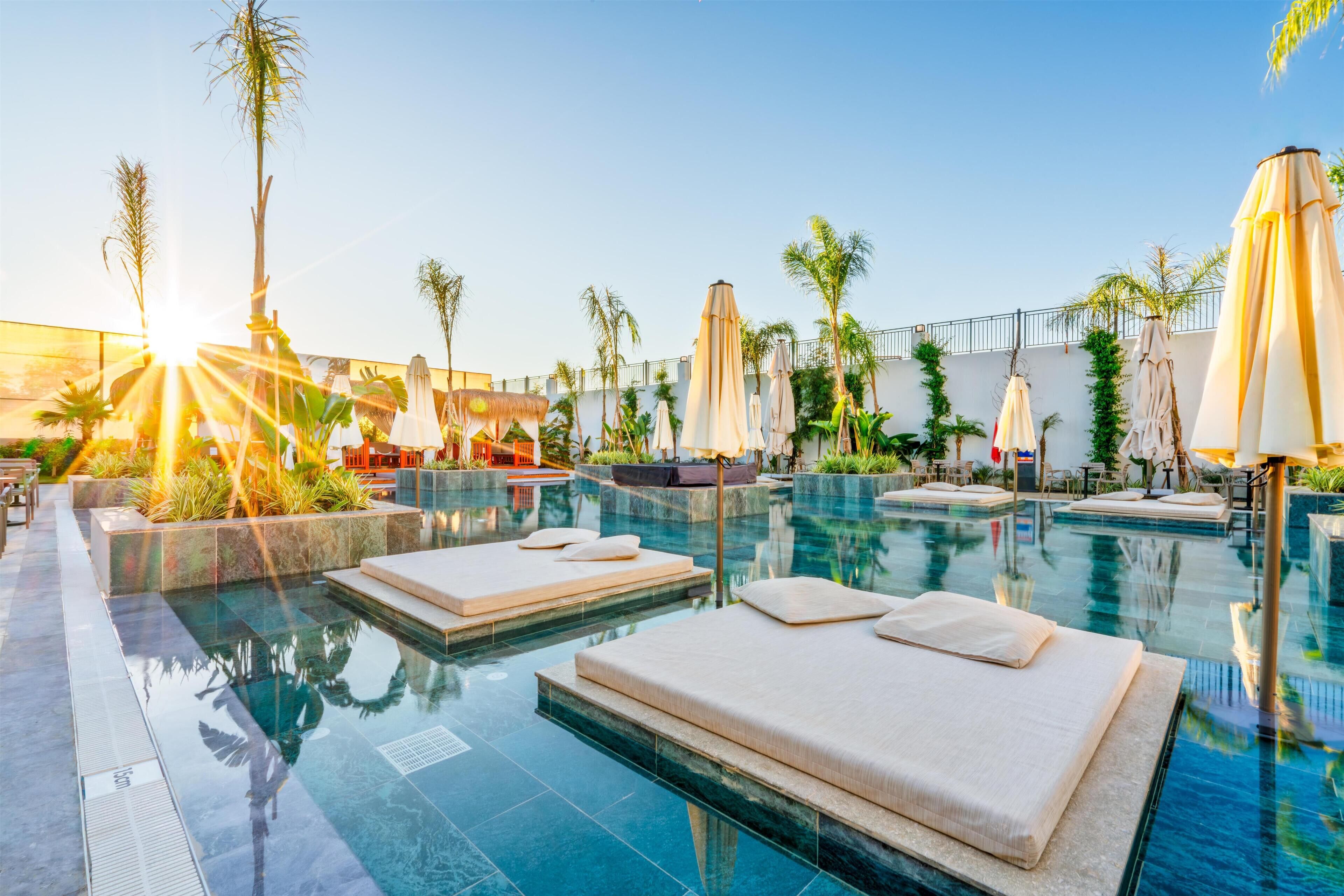 A pool area with lounge chairs and umbrellas, surrounded by palm trees and a clear sky.
