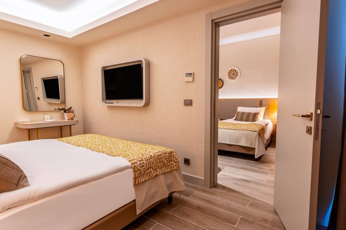 A hotel room with a bed, a TV, a mirror, a bedside table, and a door leading to another room.