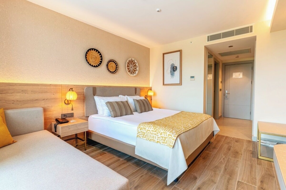 A hotel room with a large bed, two bedside tables, a nightstand, and a bedside lamp. There are decorative wall plates above the bed and a framed picture on the wall.