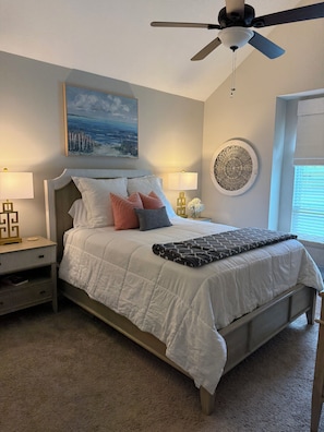 A bedroom with a bed, a ceiling fan, a painting, a bedside table with a lamp, and a decorative wall piece.