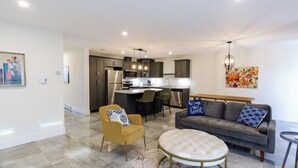 A modern kitchen with a central island, dining area with chairs, a sofa, and a round ottoman.