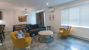 A modern living room with a sofa, two armchairs, a round ottoman, a dining table, and a ceiling fan.