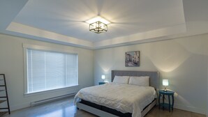 A bedroom with a bed, two bedside tables with lamps, a window with blinds, and a painting on the wall.