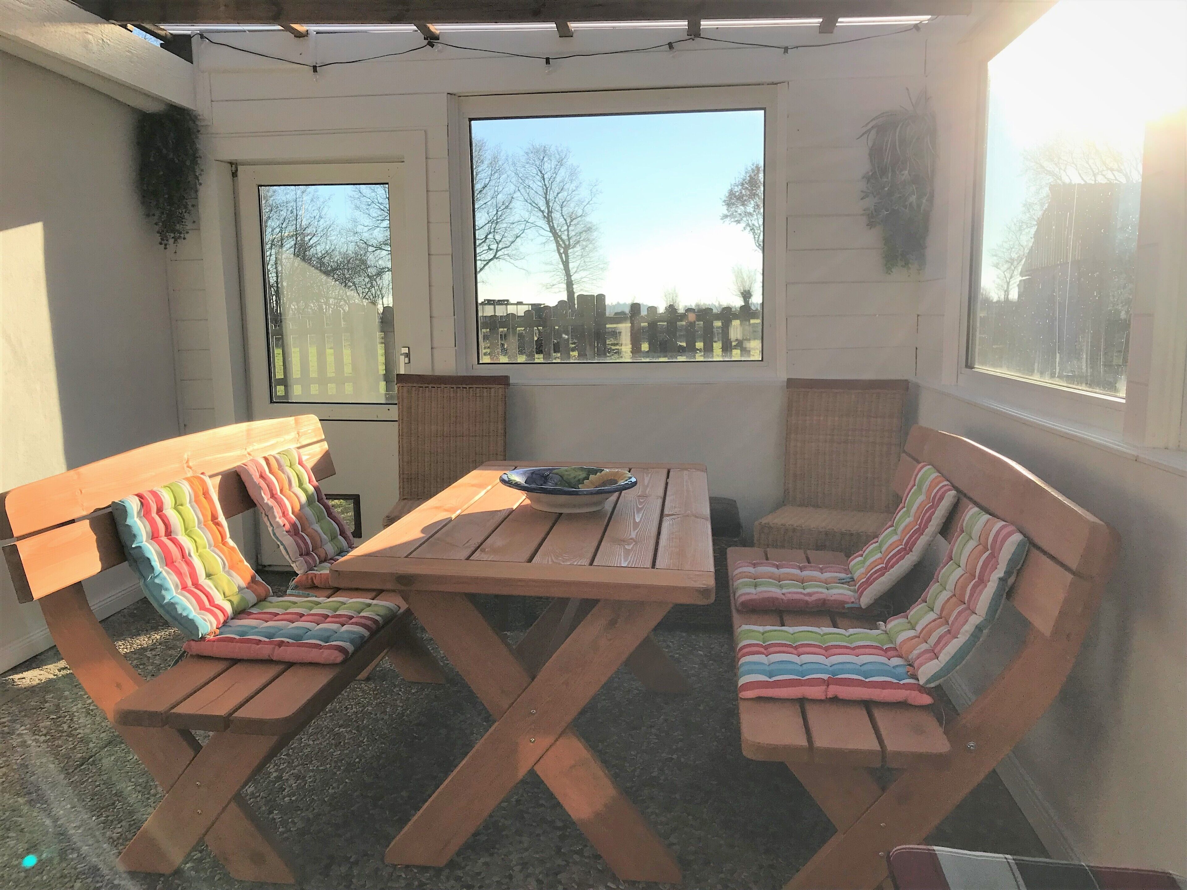 Une terrasse avec des bancs en bois et une table, entourée de fenêtres donnant sur une clôture et des arbres.