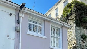A two-story house with a light pink exterior, a front door, and a small window with a plant on the second floor. - Ringmore Cottage - Salcombe - View over estuary (Salcombe)
