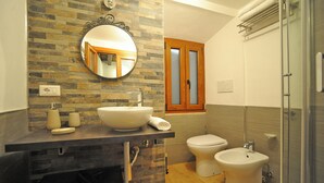 Un bagno con parete in pietra, uno specchio tondo, un lavandino, un water, un bidet e un box doccia in vetro.