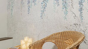 A wicker chair with a red cushion, a small table with candles, and a wall with a leaf pattern.
