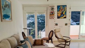 A living room with a beige sofa, a patterned ottoman, a wooden rocking chair, and a small table. There are two windows with a view of a hillside. - Stunning Central Penthouse with Breathtaking Sea and Mountain Views (Benalmádena)
