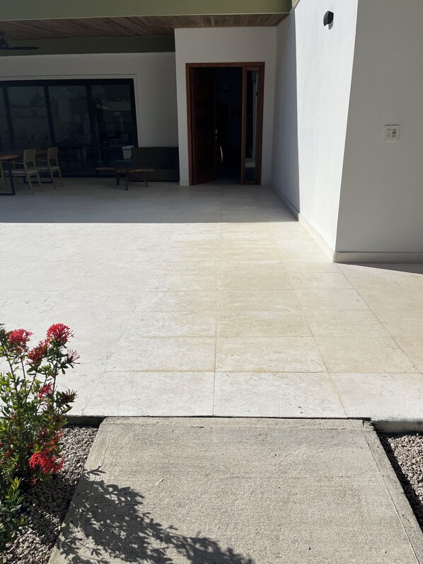 An outdoor patio area with a tiled floor, a wooden bench, and a door leading to an interior space. - Gated + Lux +New Modern 4-BD Home in Playa Potrero – AC, WiFi & Steps to Beach! (Playa Potrero)