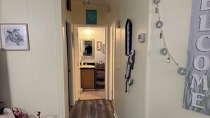Un pasillo con piso de madera, un cuadro enmarcado de una tortuga, un letrero que dice 'WELCOME' y una pequeña área de cocina con un lavamanos y armarios.