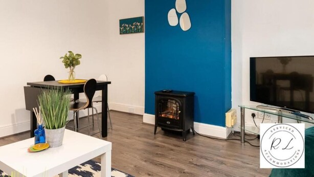 A modern living room with a blue accent wall, a dining area with chairs and a table, a black wood-burning stove, and a flat-screen TV. - 1br-2guests-freeparking-wifi-tv (Wigston)