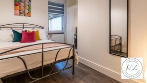 A bedroom with a bed, colorful pillows, a window with blinds, a mirror, and a wall art piece. - 1br-2guests-freeparking-wifi-tv (Wigston)