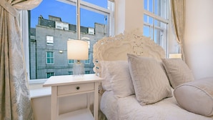 Ein Schlafzimmer mit Bett, Nachttisch, Lampe und Blick auf Gebäude außerhalb des Fensters.