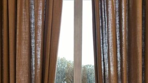A window with brown curtains, a radiator below, and a view of a building and trees outside. - Corvetto View (Genoa)