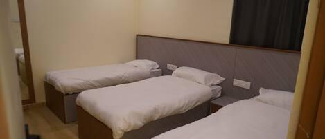 A hotel room with three single beds, a wooden headboard, a window with a dark curtain, and a light switch on the wall.