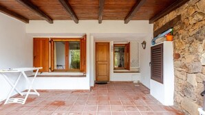 Ein überdachter Patio-Bereich mit Fliesenboden, Holzbalken, einem weißen Tisch und einer Steinwand.