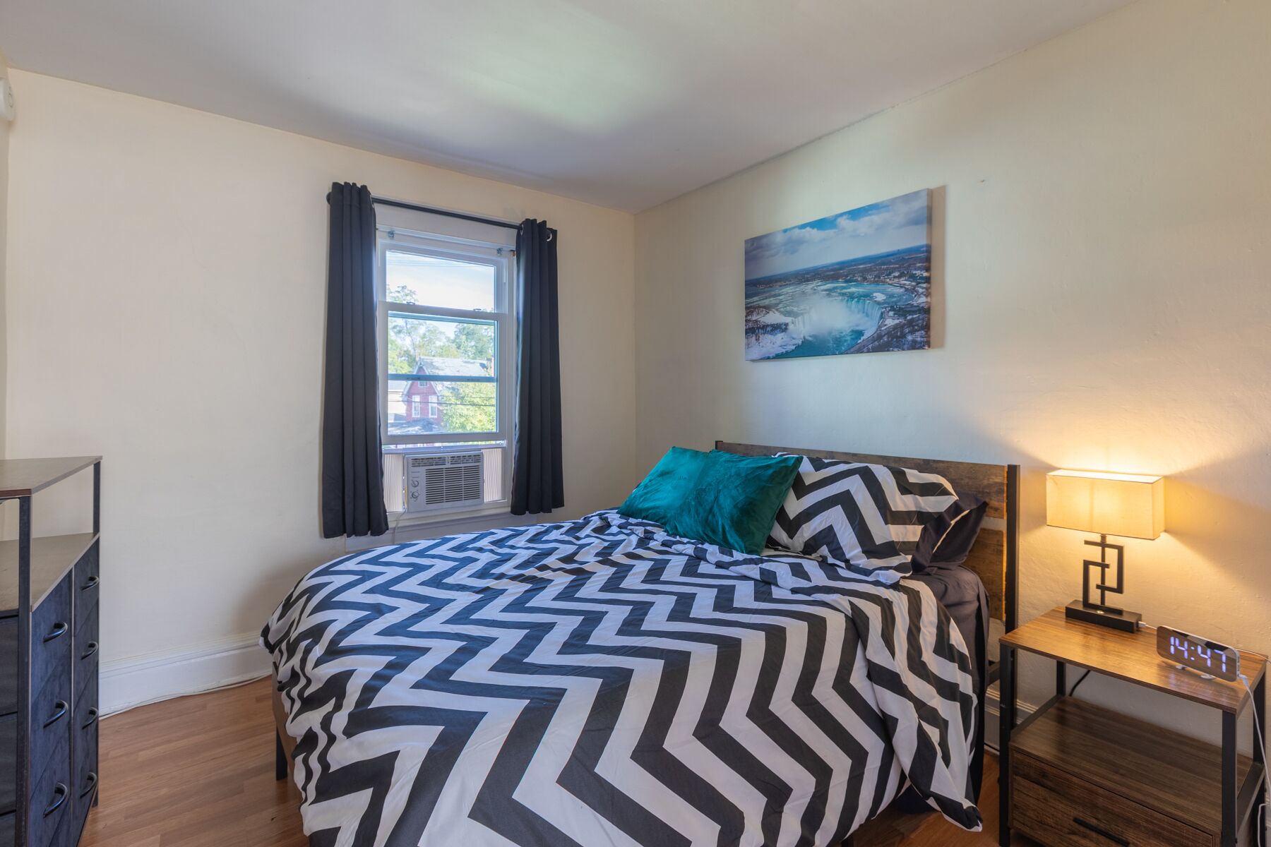 Een slaapkamer met een bed, een nachtkastje, een lamp, een klok, een raam met gordijnen en een schilderij aan de muur.