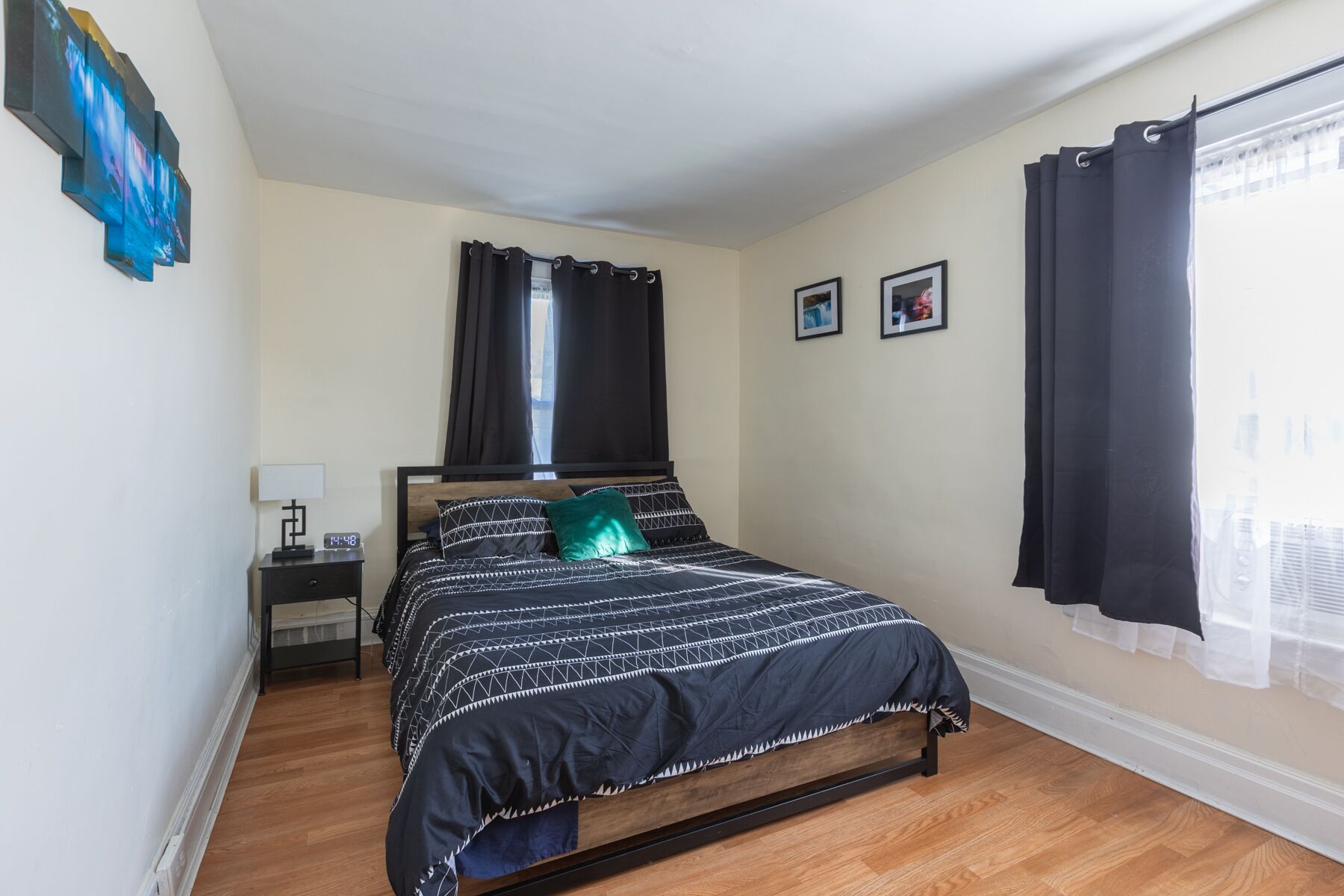 A bedroom with a bed, a nightstand, a window with curtains, and a wall-mounted artwork.