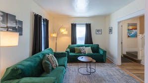 A living room with two green sofas, a small round table, and a floor lamp. There are curtains on the windows, and a poster on the wall.