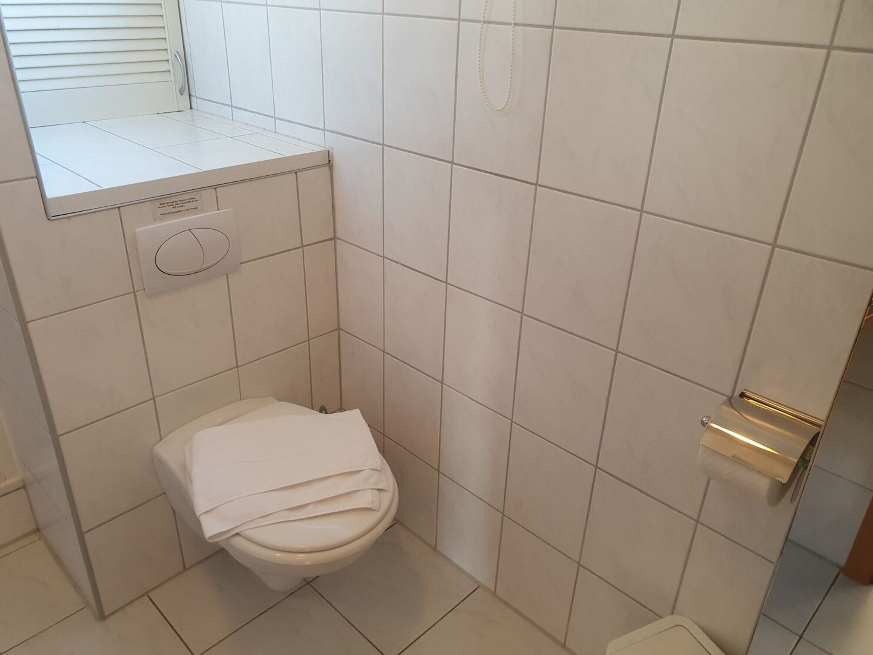 A small, compact bathroom with white tiles, a toilet, a towel rack, and a small window with blinds.