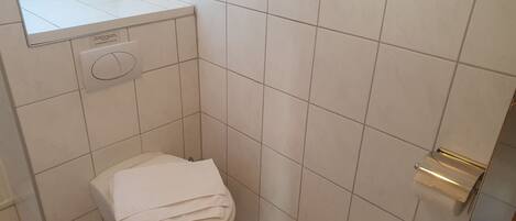 A small, compact bathroom with white tiles, a toilet, a towel rack, and a small window with blinds.