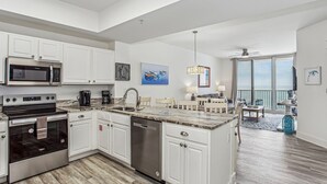 A modern kitchen with white cabinetry, a built-in microwave, stove, and dishwasher. There is a dining area with a table and chairs, and a balcony with a view. - Absolutely Shore, Navarre Beach (Navarre)