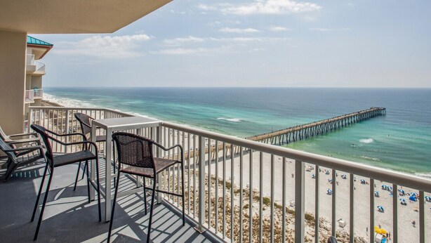 A balcony with two wicker chairs, a view of the ocean, a pier, and a beach with umbrellas. - Absolutely Shore, Navarre Beach (Navarre)