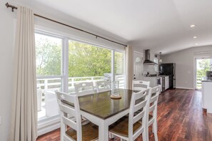 Área de refeições com mesa e cadeiras, cozinha equipada e grandes janelas com vista para o verde.