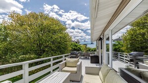 Une terrasse extérieure couverte, aménagée avec des meubles en osier, un barbecue et offrant une vue sur une cour bordée d’arbres.